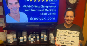 Smiling woman, in Santa Clarita Chiropractor office, holding hand written sign that says “1 year of bad TMJ 6 months of specialist, no better. 4 treatments here 90% better!”