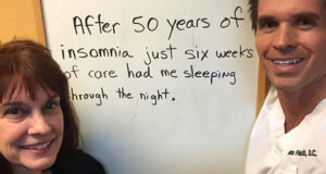 Smiling woman standing next to Santa Clarita chiropractor Dr. Thomas Polucki with a handwritten sign that says: “After 50 years of insomnia, just six weeks of care had me sleeping through the night.”